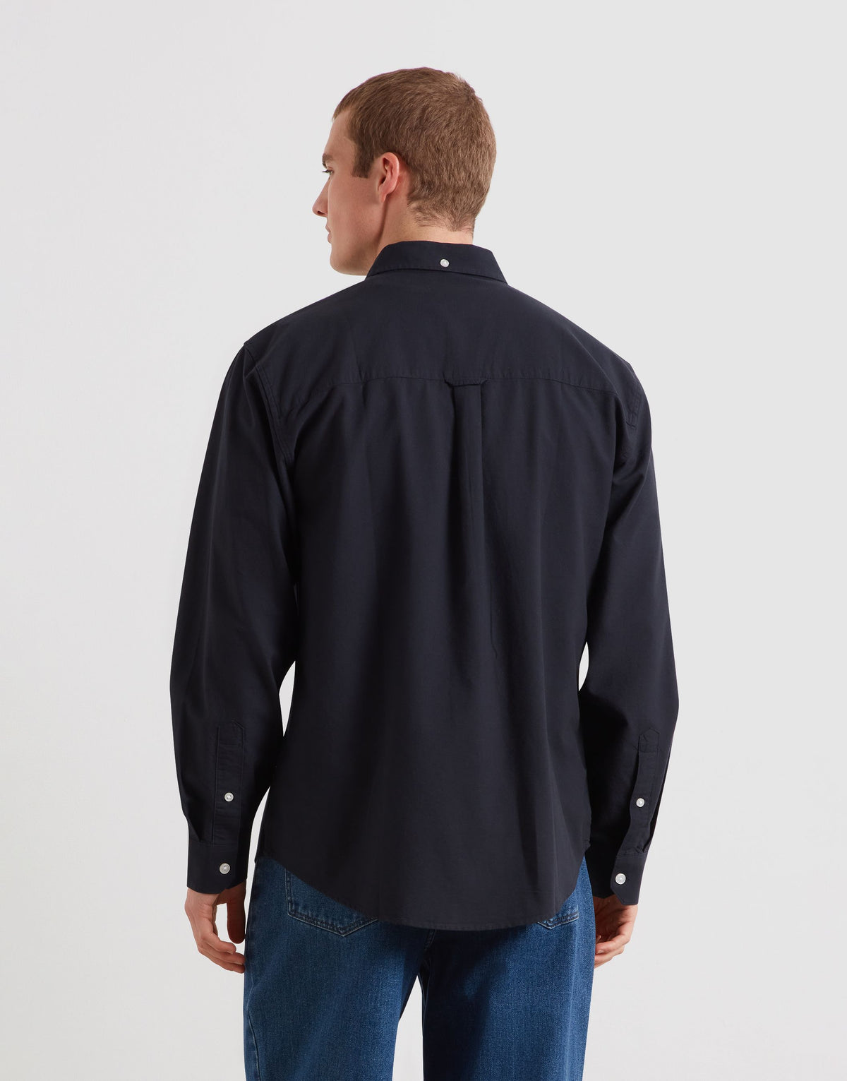 A man viewed from the back wearing a dark navy long-sleeve button-up shirt and blue jeans against a light gray studio background.