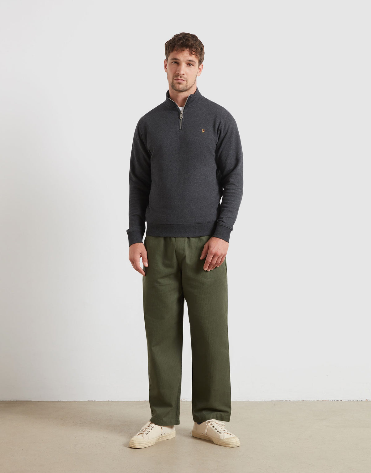 A man modeling a dark charcoal gray quarter-zip pullover sweater with a small embroidered Farah gold F logo on the chest, paired with olive green trousers and off-white sneakers, standing against a plain light gray studio background.