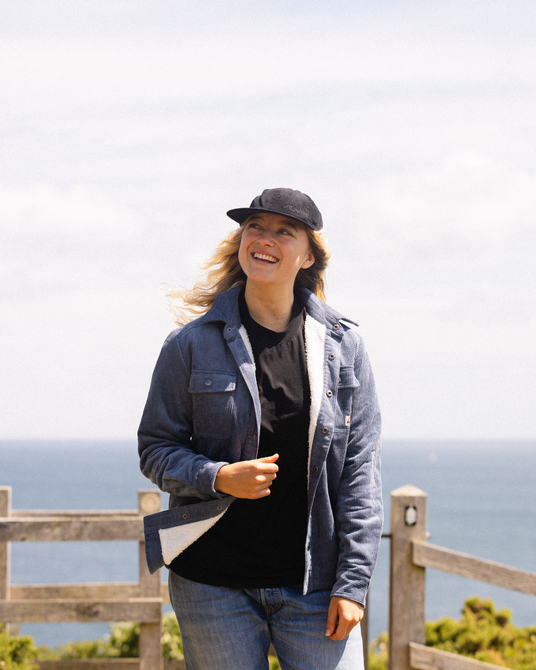 A woman smiling outdoors by the ocean, wearing a dark blue denim jacket over a black t-shirt and blue jeans, with a black baseball cap. A wooden fence and the sea are visible in the background.