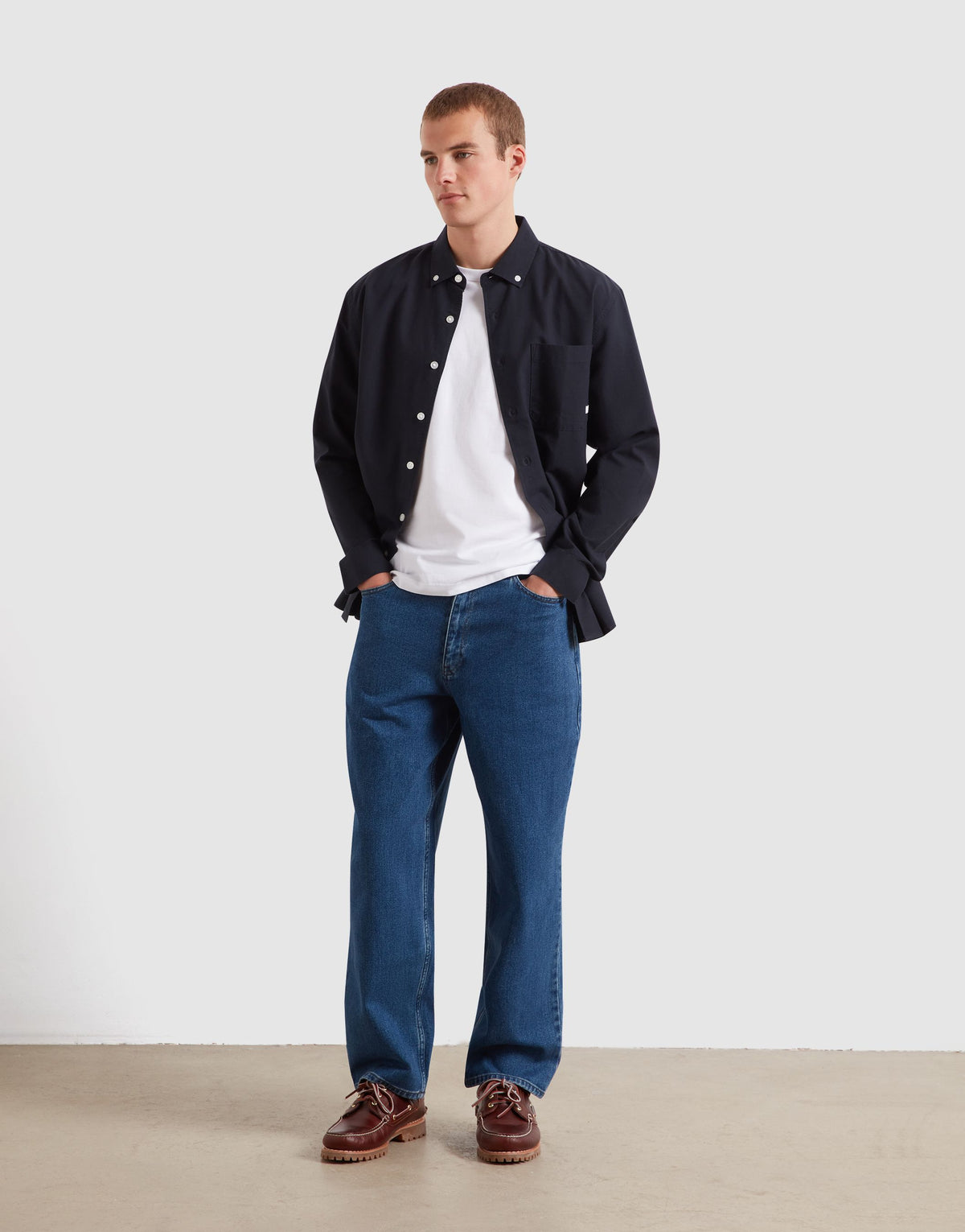 A man modeling a dark navy overshirt over a white t-shirt, paired with blue jeans and brown leather shoes, standing against a plain light gray studio background.