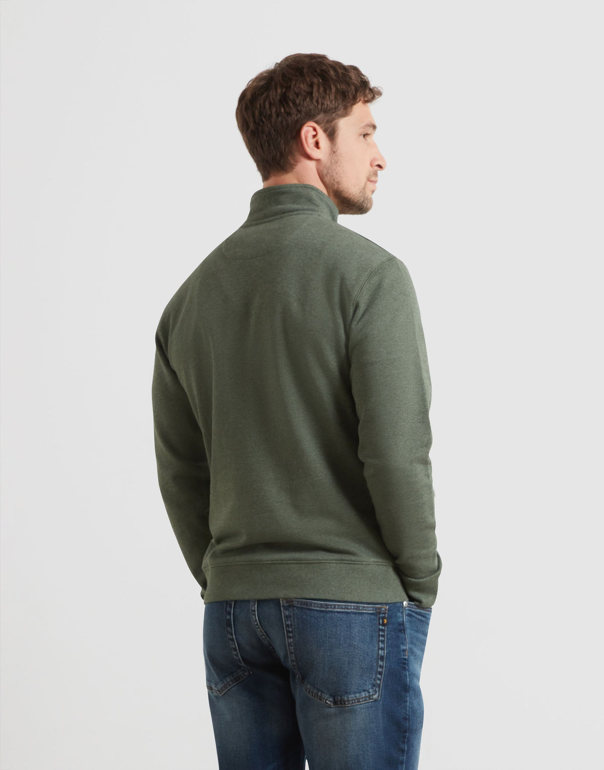 A man wearing an olive green quarter-zip pullover sweater and blue jeans, shown from a three-quarters back view against a light gray studio background.
