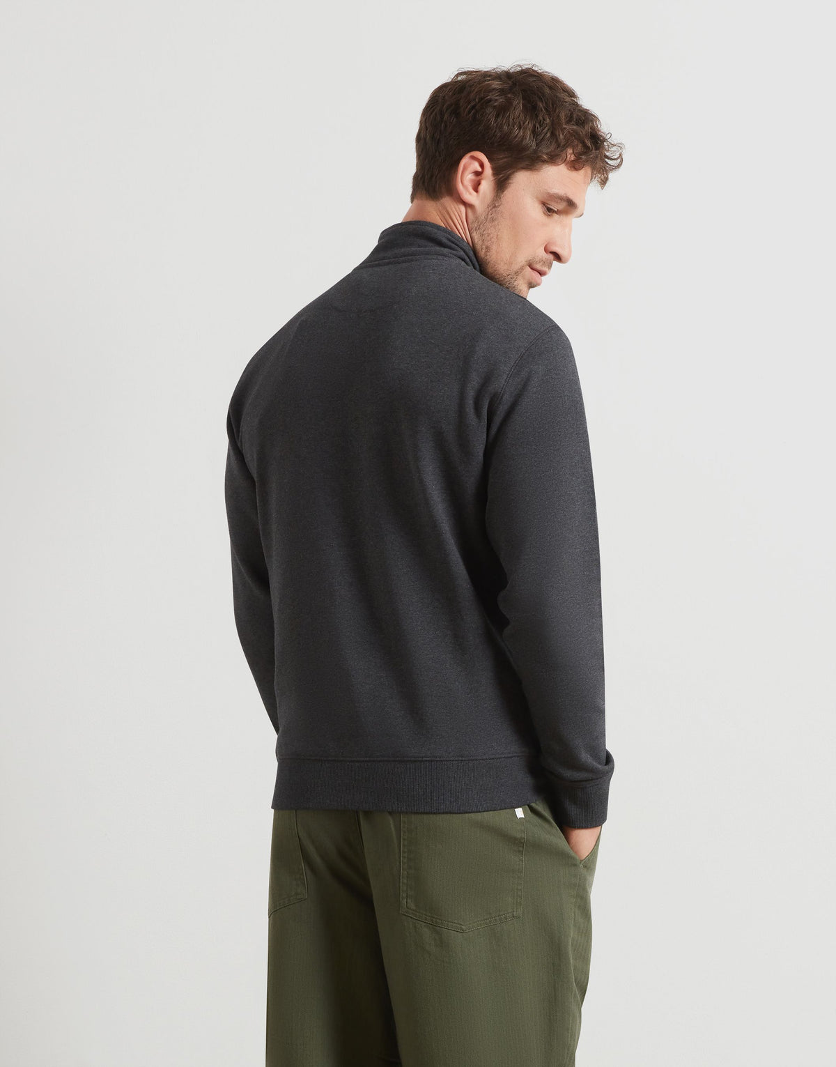 A man wearing a dark charcoal gray quarter-zip pullover sweater and olive green trousers, shown from a three-quarters back view against a light gray studio background.