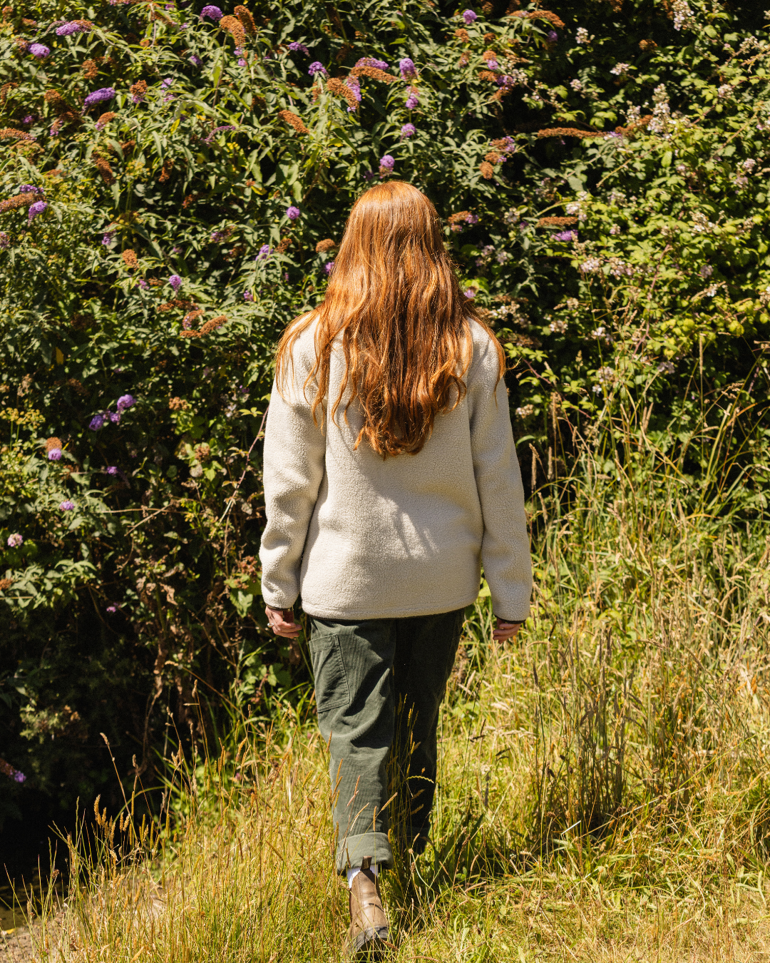 A woman with long red hair is seen from behind, walking away in a grassy field with dense green bushes and purple flowers in the background. She is wearing a cream-colored fleece, dark green pants, and brown boots.