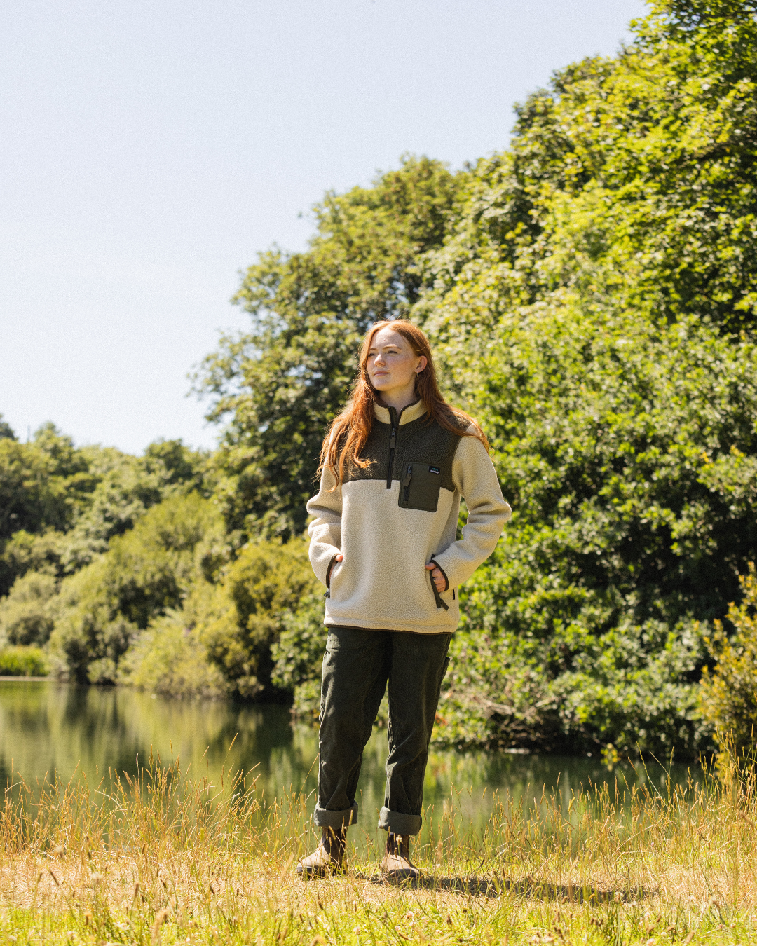 A woman stands outdoors by a lake with trees in the background, wearing a light beige and dark green quarter-zip fleece pullover, dark green pants, and brown hiking boots.