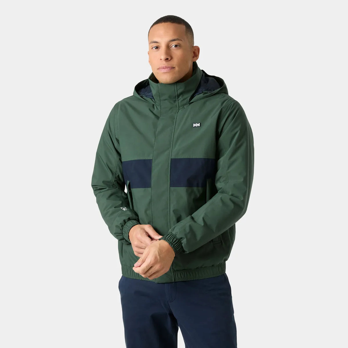A man wearing a green and navy hooded jacket with a small white Helly Hansen logo on the chest, paired with navy pants, against a light gray studio background.