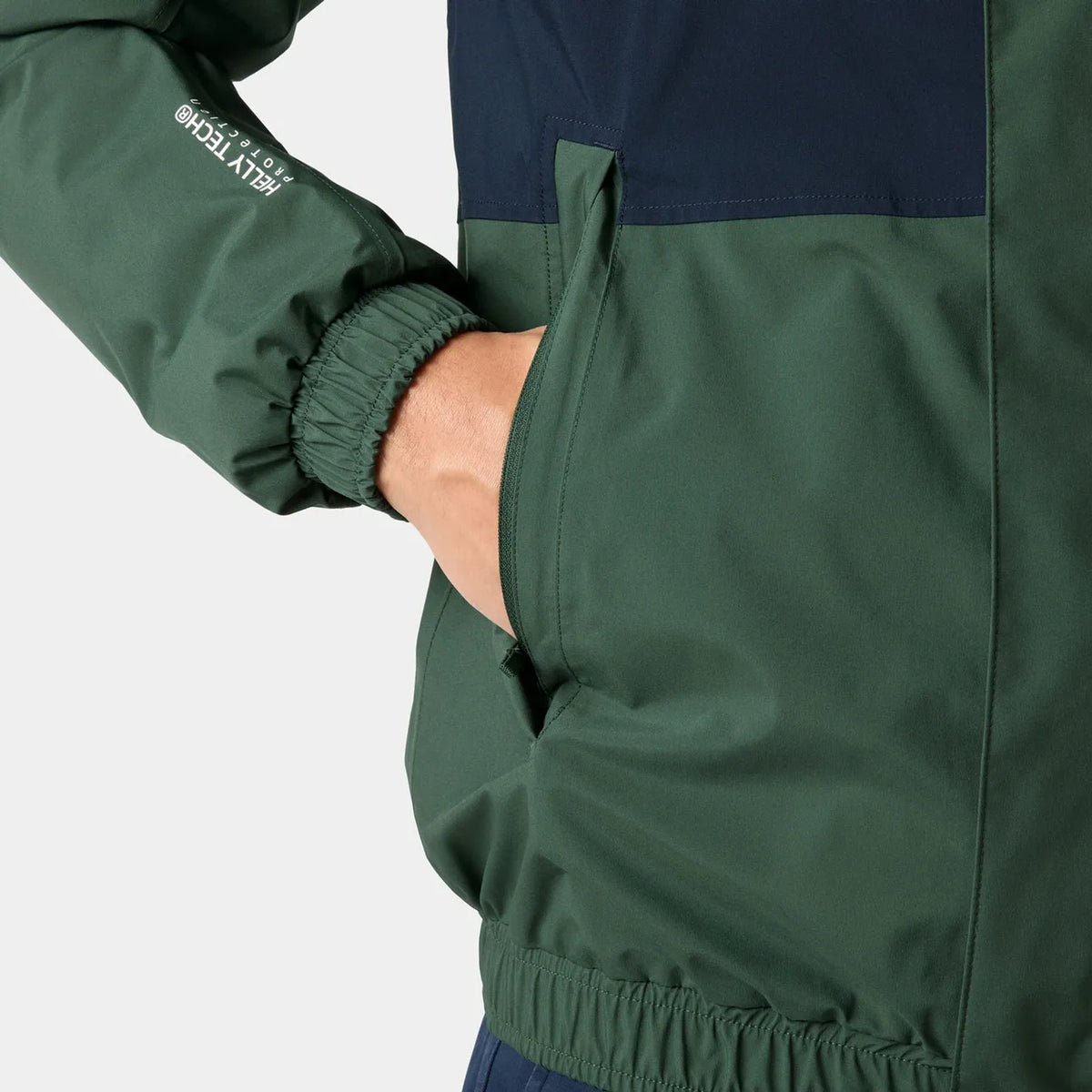 Close-up of a person wearing a dark green and navy colorblock jacket, showing the sleeve with a white 'HELLY TECH' logo and a side pocket with the hand inside.