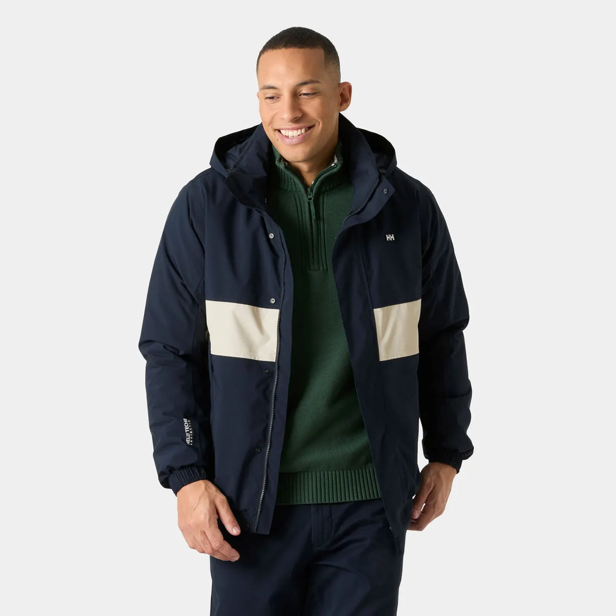 A man wearing a navy blue hooded jacket with beige color-blocked panels on the chest, layered over a dark green quarter-zip sweater, against a light gray studio background.