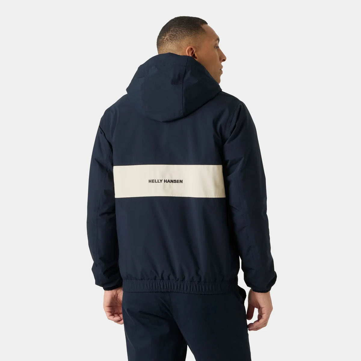A man shown from the back wearing a navy blue hooded windbreaker jacket with a wide cream horizontal stripe across the upper back and the brand name 'HRLLY HANSEN' printed in navy. He is also wearing matching navy pants against a light gray studio background.