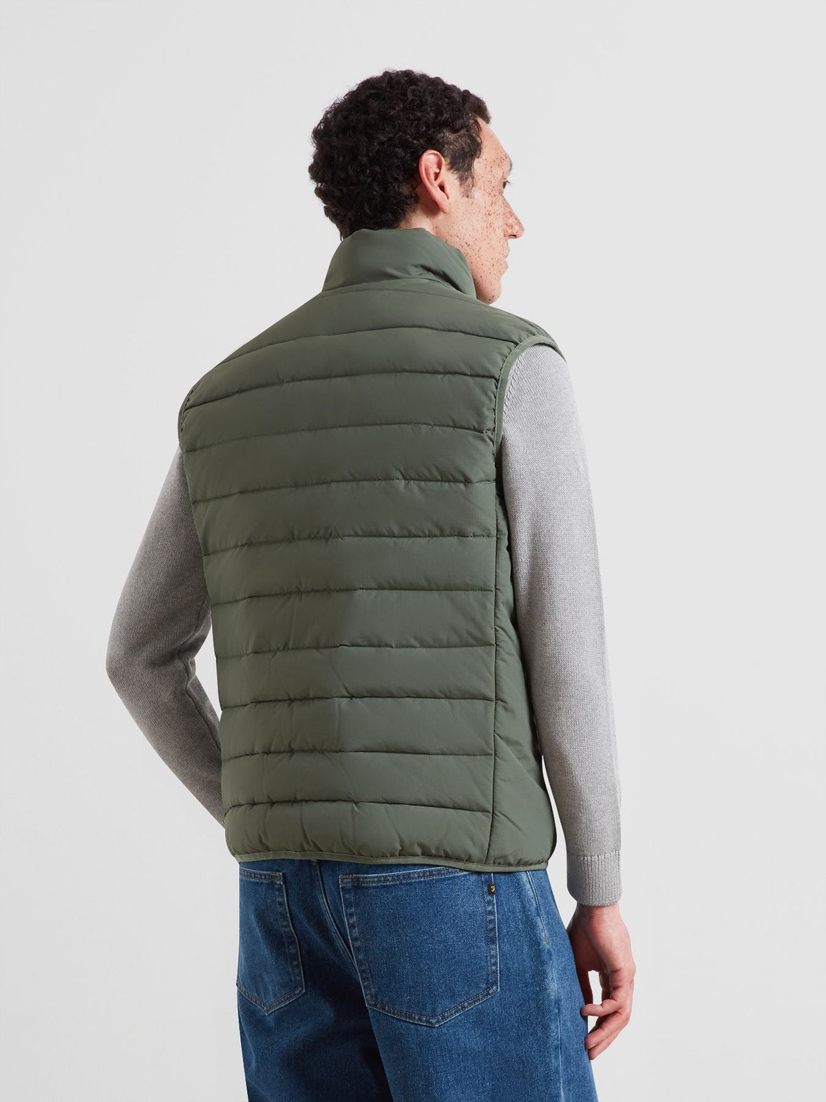 A man shown from the back wearing an olive green puffer vest over a gray long-sleeve shirt and blue jeans, photographed against a light gray studio background.