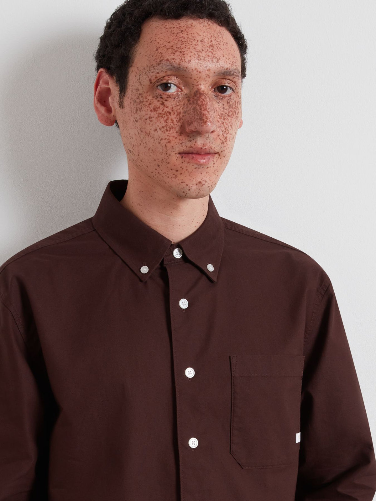 A man with freckles wearing a dark brown button-down shirt with white buttons, photographed against a light gray wall.