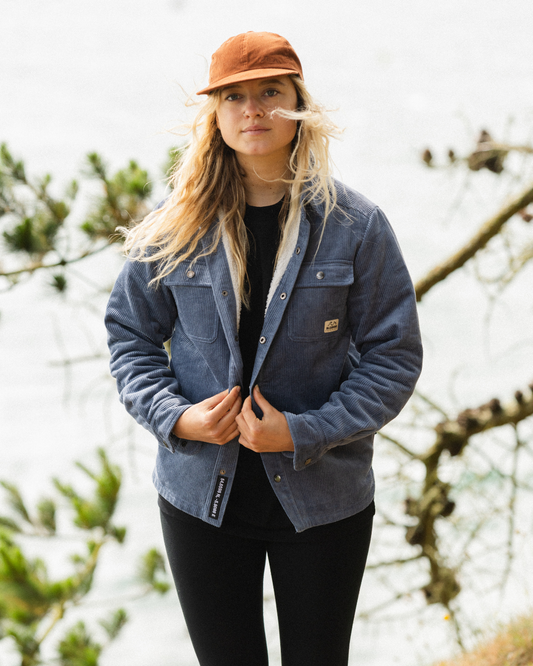 A woman wearing a blue denim-style button-front overshirt jacket, black top and pants, and an orange cap, standing outdoors with blurred tree branches and foliage in the background.