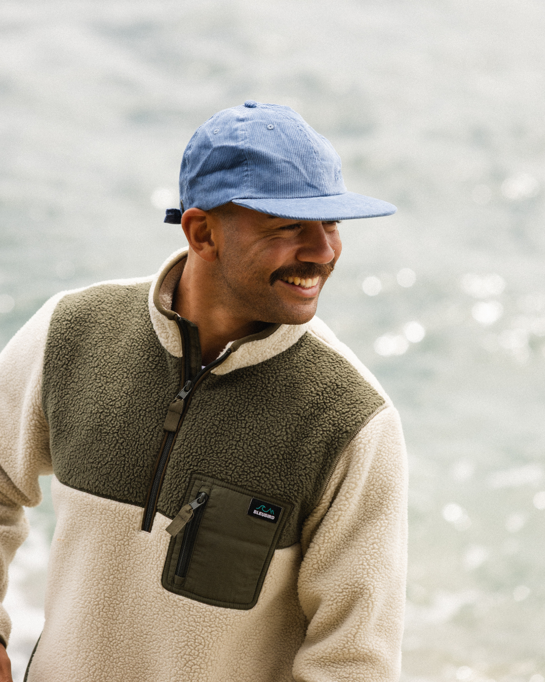 A man wearing a Bleubird fleece pullover in beige and olive green with a blue cap, smiling outdoors with a blurred natural background.