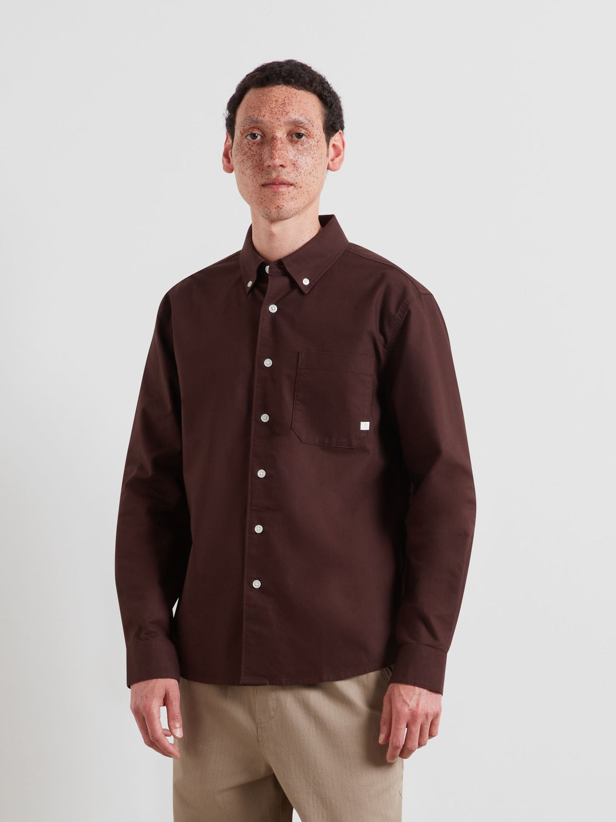 A man wearing a dark brown long-sleeve button-down shirt with a chest pocket and beige pants, standing against a light gray studio background.