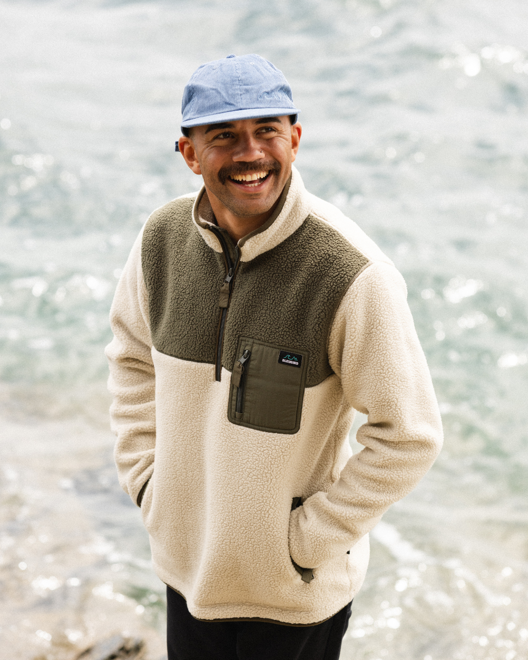 A man smiling outdoors by the water, wearing a Bleubird quarter-zip fleece pullover in beige and olive green with a blue cap.