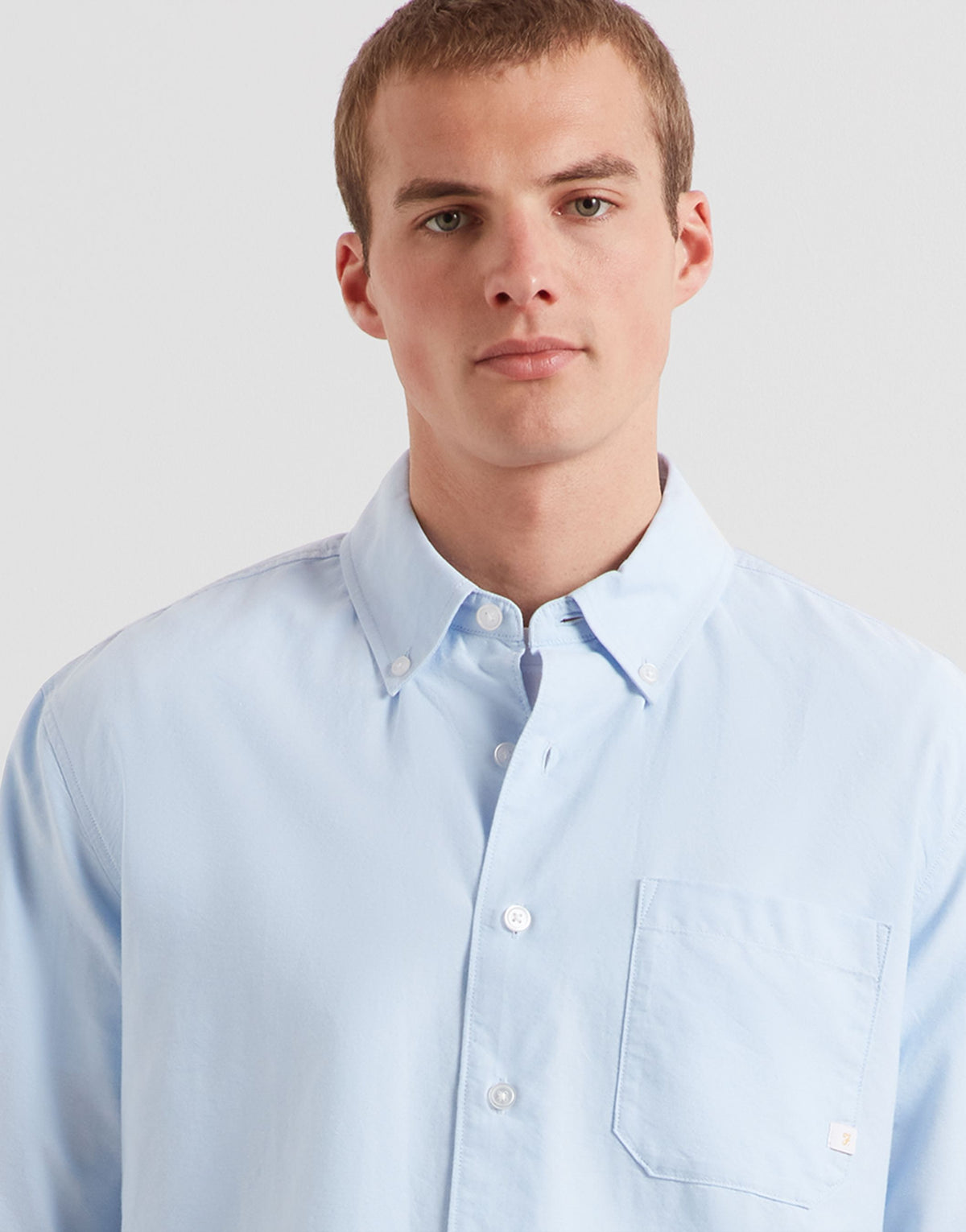A man wearing a light blue long-sleeve button-down shirt with a chest pocket, photographed against a plain light gray studio background.