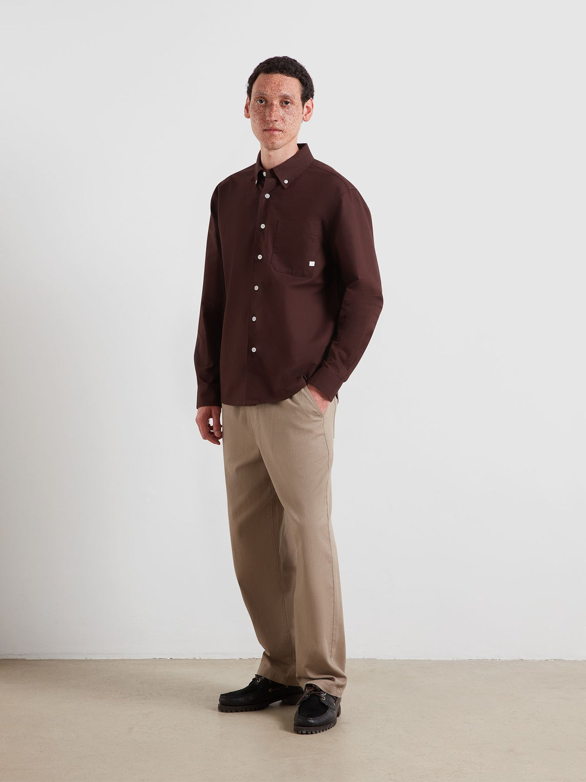 A man wearing a dark brown long-sleeve button-up shirt with a chest pocket, paired with beige trousers and black shoes, standing against a plain light gray studio wall.