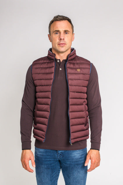 Tommy Bowe wearing a burgundy quilted puffer vest over a dark brown long-sleeve polo shirt and blue jeans, standing against a light gray studio background.
