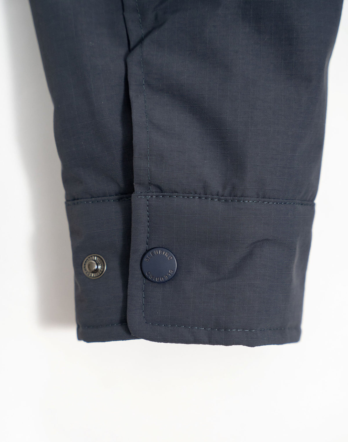 Close-up of a dark navy sleeve cuff with two buttons, one metallic and one navy blue with 'BLEUBIRD' printed on it, shown against a plain white background.