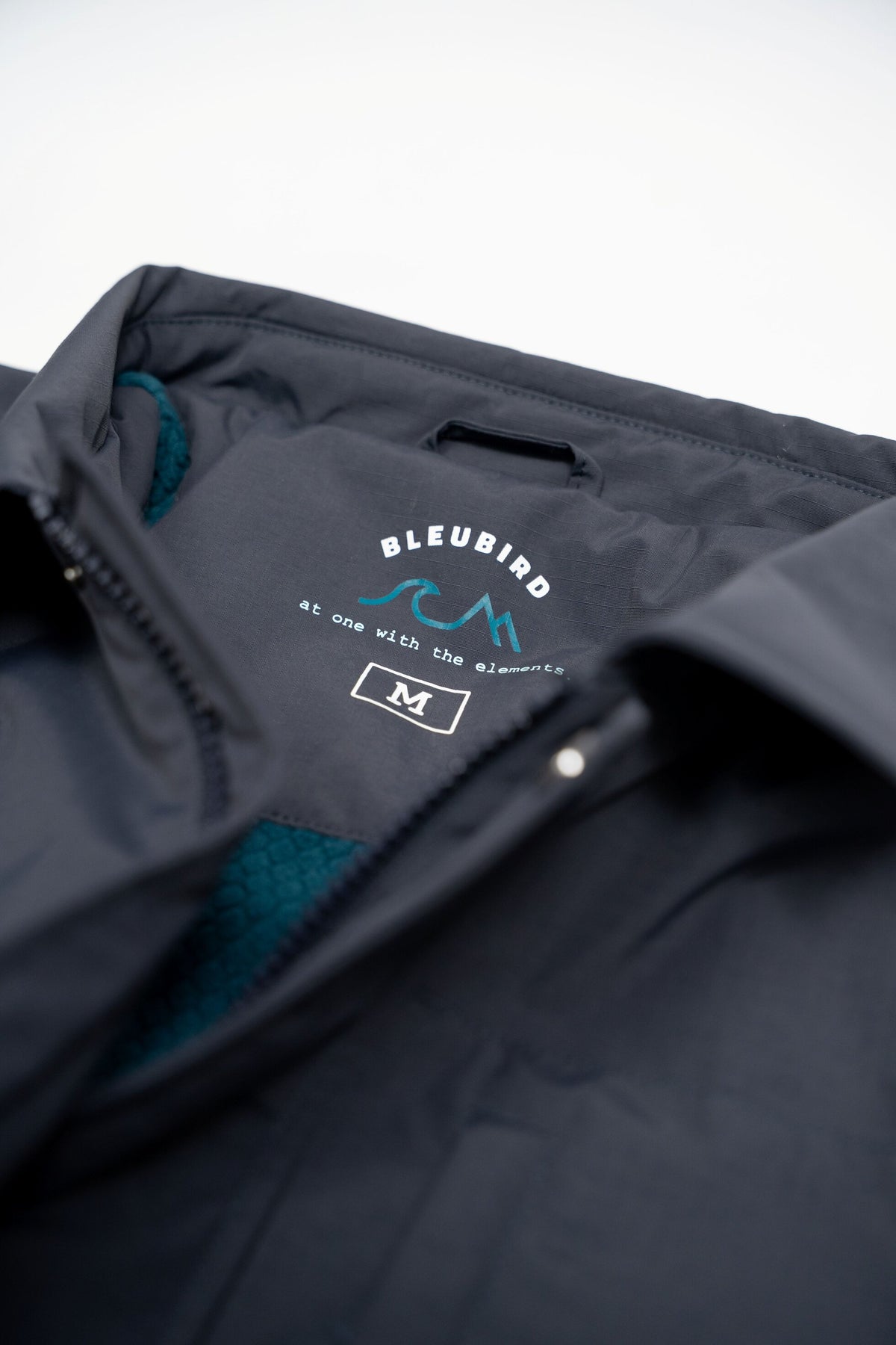 Close-up of a dark navy zippered jacket with a visible inner neck label reading BLEUBIRD and a size mark of M, with a teal textured lining visible at the collar.
