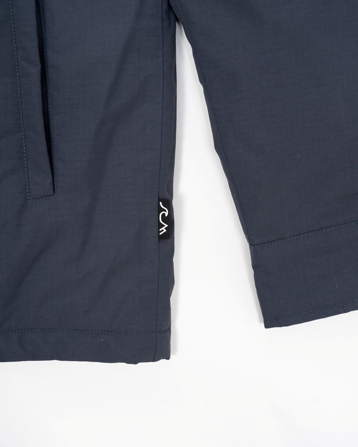 Close-up of dark slate blue jacket sleeve showing a side pocket with a small white embroidered logo, shot against a plain white background
