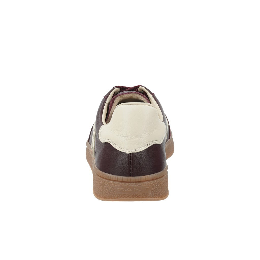 A single low-top sneaker shown from the back, featuring dark brown leather panels, a cream-colored heel counter, and a brown textured rubber sole, set against a plain white background.