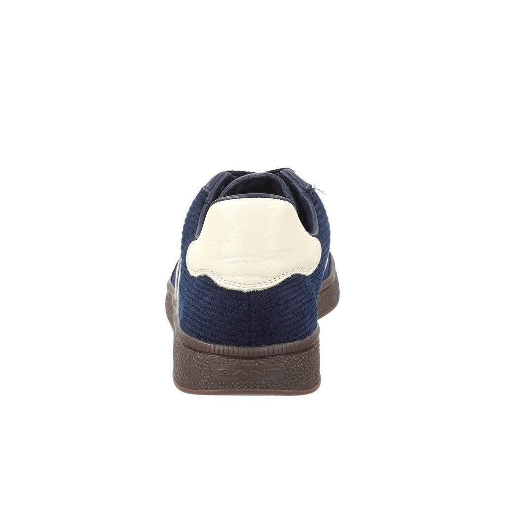A single low-top sneaker shown from the back, featuring a navy blue corduroy upper, a cream-colored heel counter, and a brown textured sole.