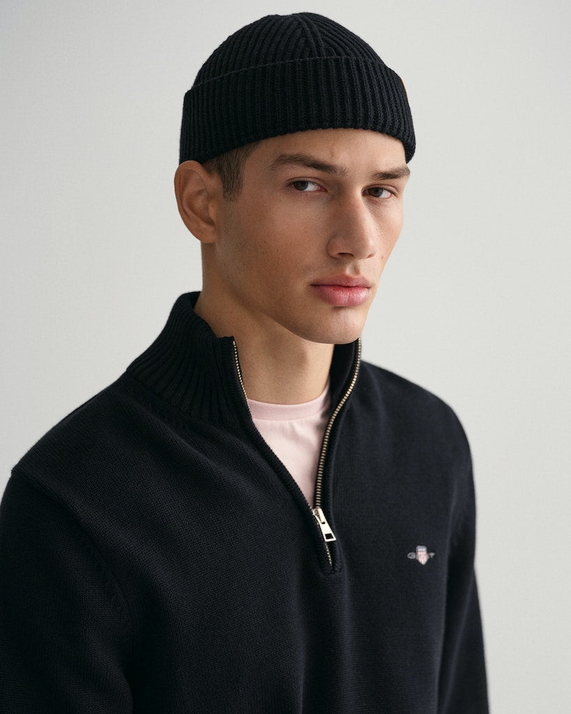 Man wearing a black knit beanie and black quarter-zip sweater on a gray background
