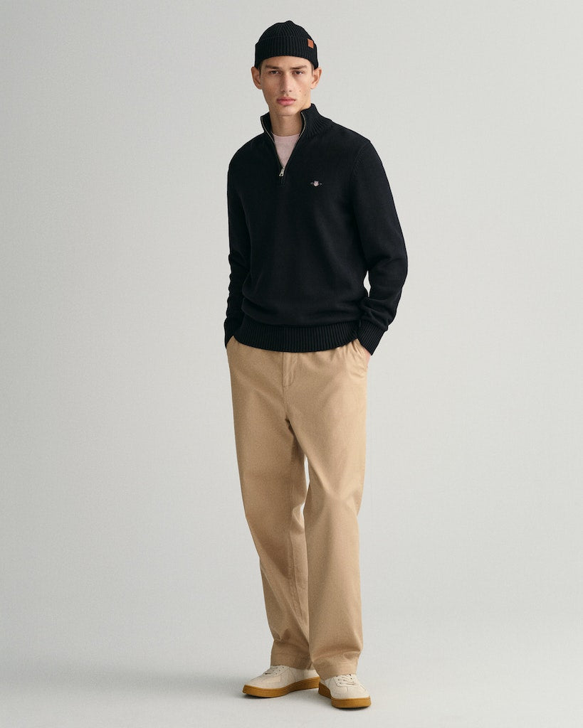 Man wearing a black sweater, beige pants, and a black cap on a light gray background