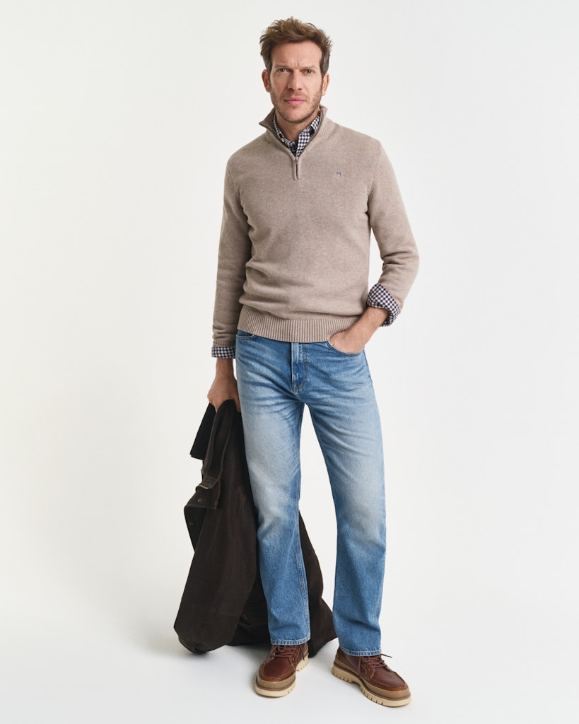 Man wearing a beige sweater, blue jeans, and brown shoes on a white background