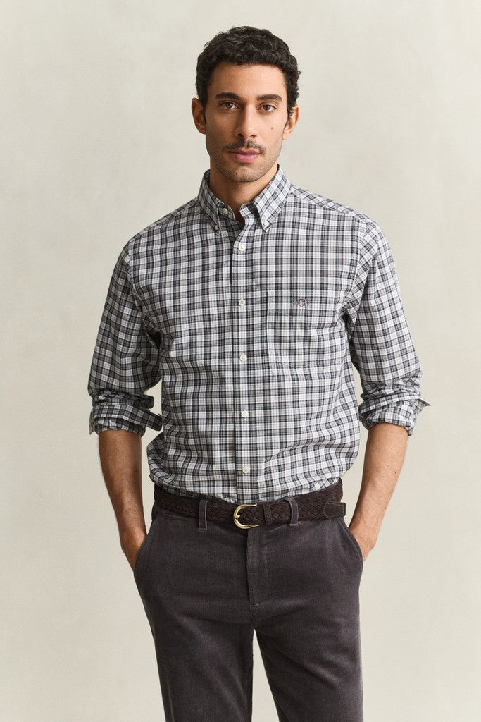 A man wearing a long-sleeve button-down shirt with a gray, white, and black plaid pattern, paired with dark gray trousers and a brown belt, standing against a light beige background.