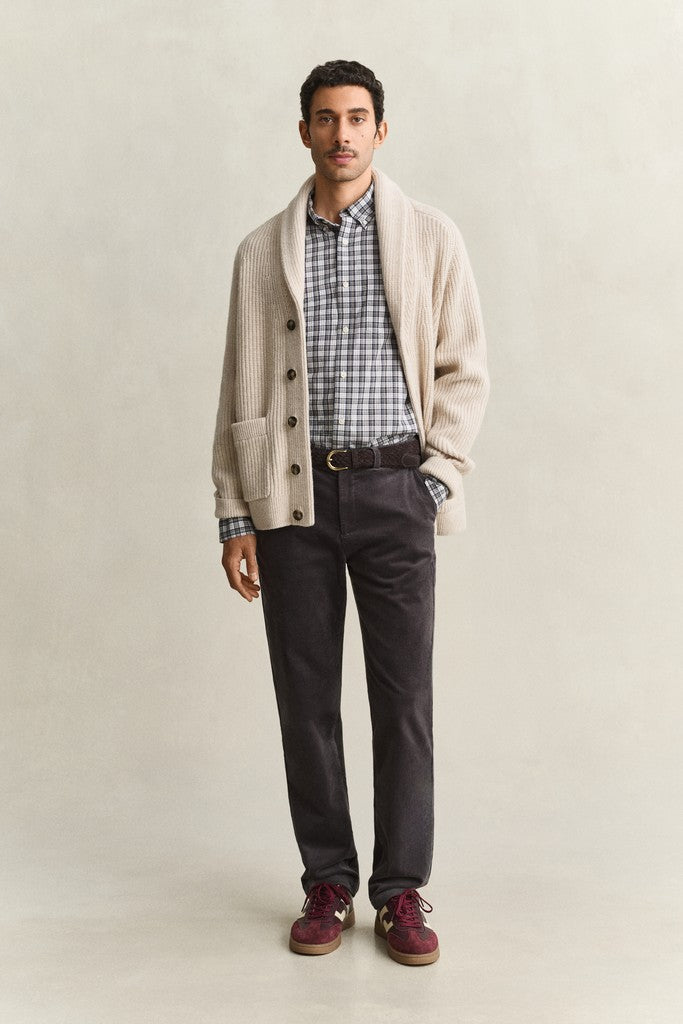 A man modeling a beige knit button-front cardigan over a blue and white plaid shirt, paired with dark charcoal trousers, a dark belt, and burgundy lace-up shoes, against a light beige studio background.