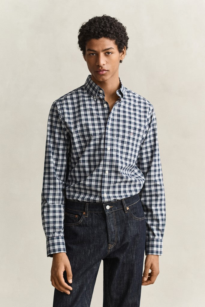 A man wearing a navy and white gingham long-sleeve button-down shirt tucked into dark blue jeans, standing against a light beige textured studio background.