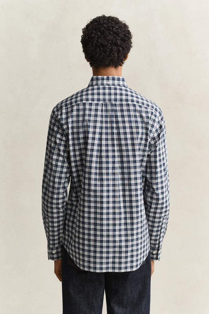 A man viewed from behind wearing a navy and white gingham plaid long-sleeve button-down shirt and dark jeans, standing against a neutral beige background.