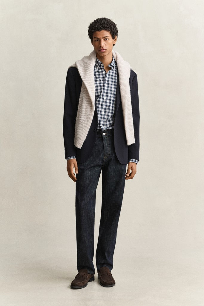 A full-length shot of a male model wearing a dark navy blazer with a cream-colored sweater, a blue and white checkered shirt, dark blue jeans, and brown suede shoes, set against a plain light beige studio background.