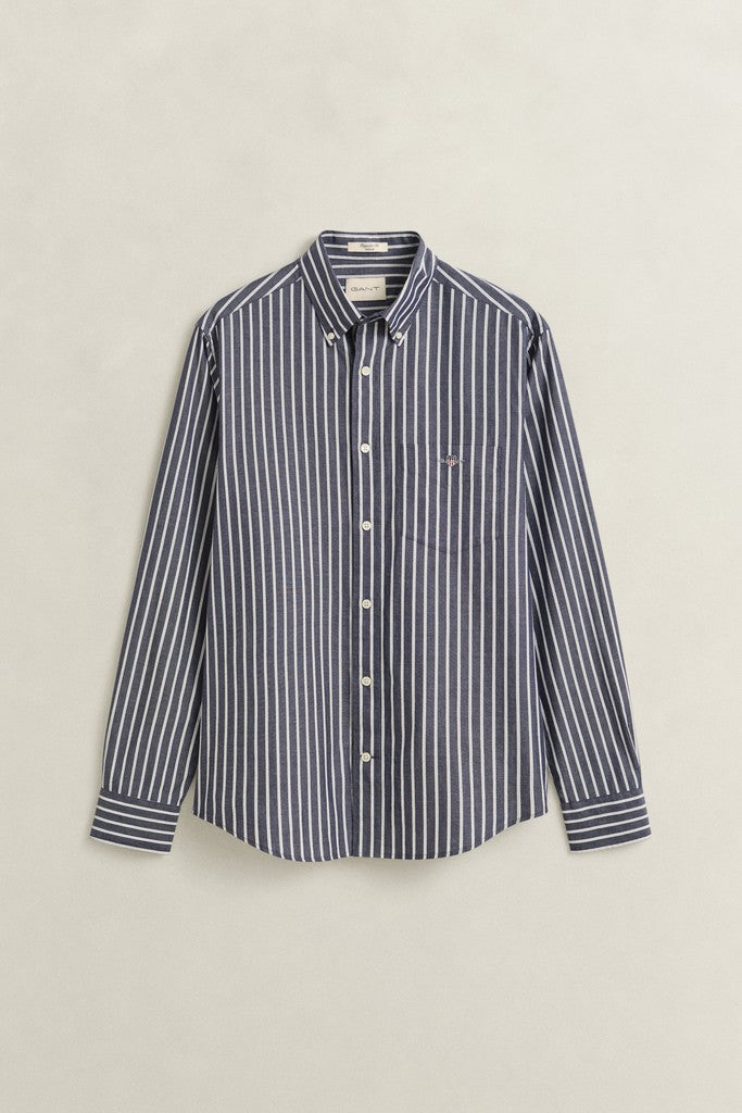 A long-sleeve button-down shirt with vertical navy and white stripes, featuring a small embroidered GANT logo on the left chest, displayed against a light beige background.