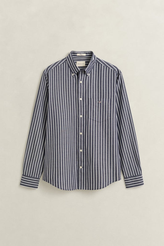 A long-sleeve button-down shirt with vertical navy and white stripes, featuring a small embroidered GANT logo on the left chest, displayed against a light beige background.