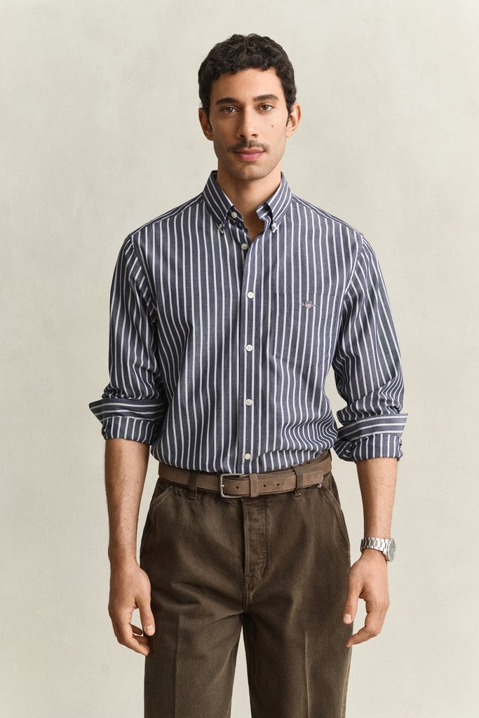 A man wearing a navy and white vertically striped button-down shirt with brown trousers, standing against a light beige studio background.