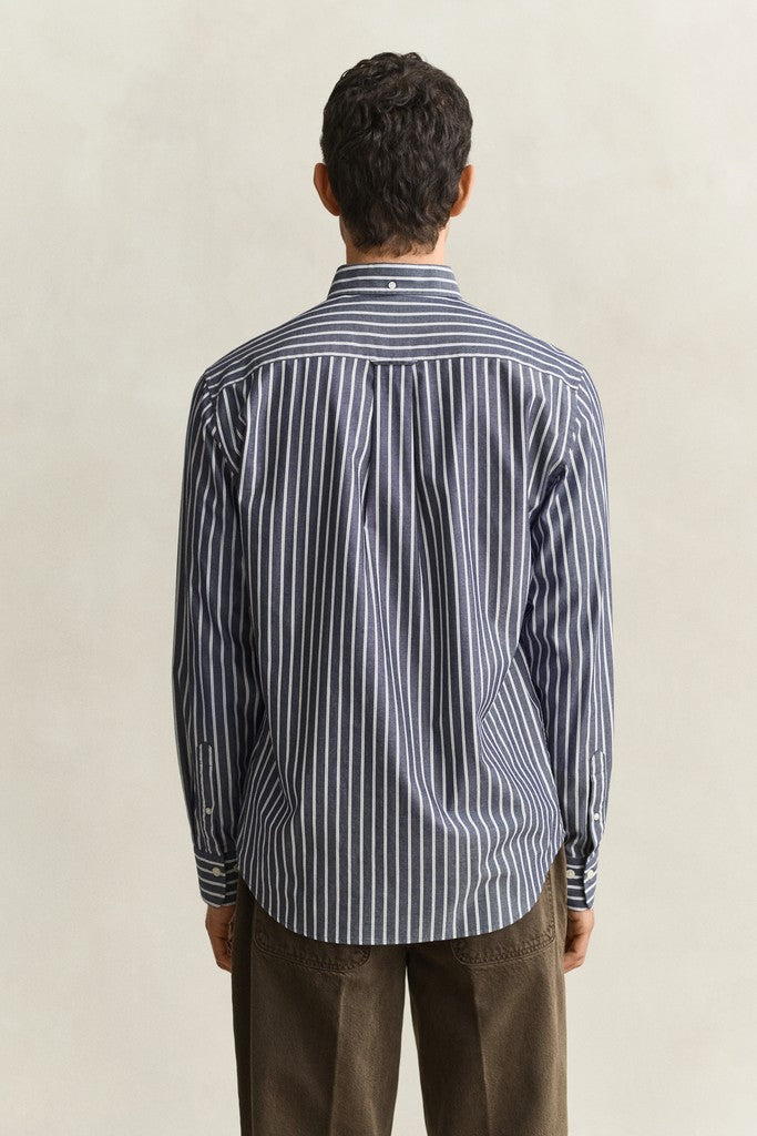 A man viewed from behind wearing a navy blue and white vertically striped long-sleeve button-down shirt and brown trousers, standing against a neutral light beige background.