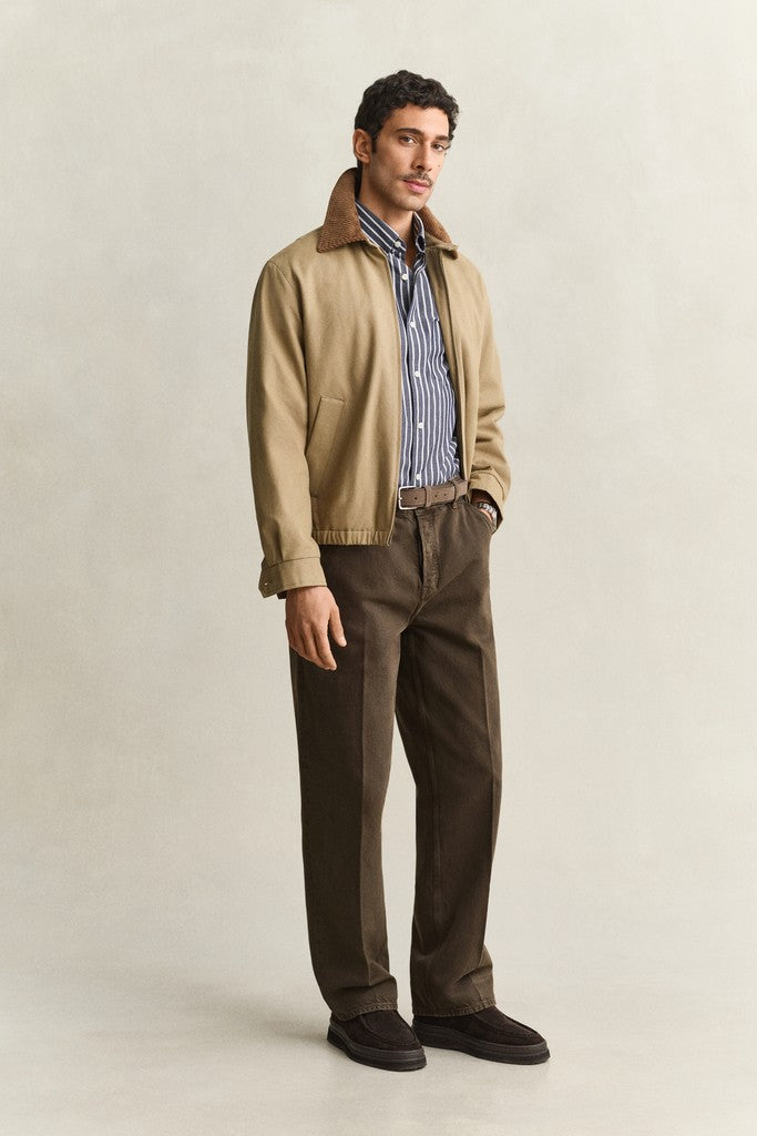 A man stands against a plain light beige studio background wearing a tan overshirt with a brown corduroy collar, a blue and white striped button-down shirt, dark brown trousers, and dark brown shoes.