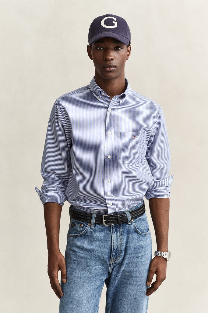 A man wearing a light blue and white vertically striped button-down shirt, blue jeans, a black belt, and a navy baseball cap with a white 'G' logo, standing against a neutral beige background.