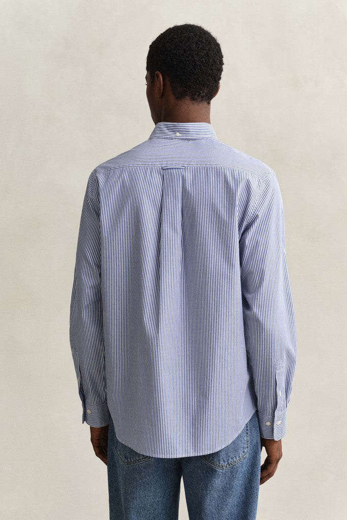 A man viewed from behind wearing a light blue and white vertically striped long-sleeve button-down shirt with blue jeans, set against a neutral beige background.