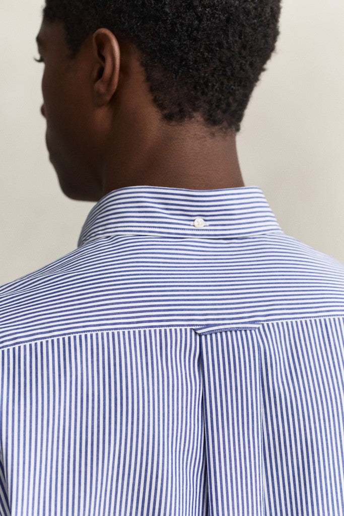 Close-up back view of a man wearing a blue and white vertically striped button-down shirt, showing the collar and upper back against a plain light beige background.
