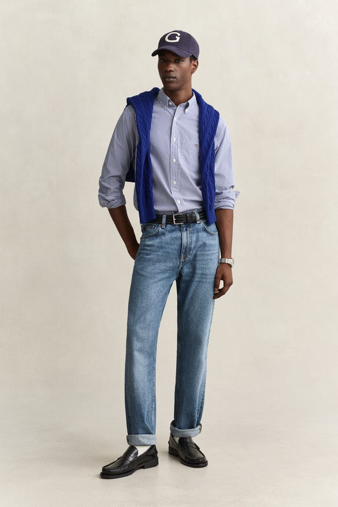 A man modeling a light blue button-down shirt, a blue knit jumper, light-wash blue jeans, black leather loafers, and a navy baseball cap with a white letter 'G' on the front, standing against a plain beige studio background.