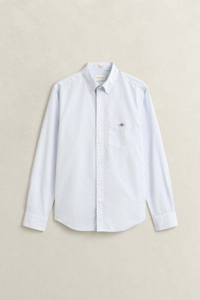 A light blue long-sleeve button-down shirt with a classic collar and a small embroidered GANT logo on the left chest, displayed flat against a neutral beige background.
