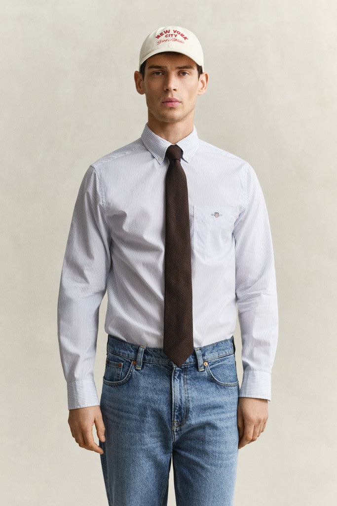 A man wearing a light blue long-sleeve button-down shirt, a dark brown tie, blue jeans, and a white baseball cap with red text, standing against a neutral beige background.