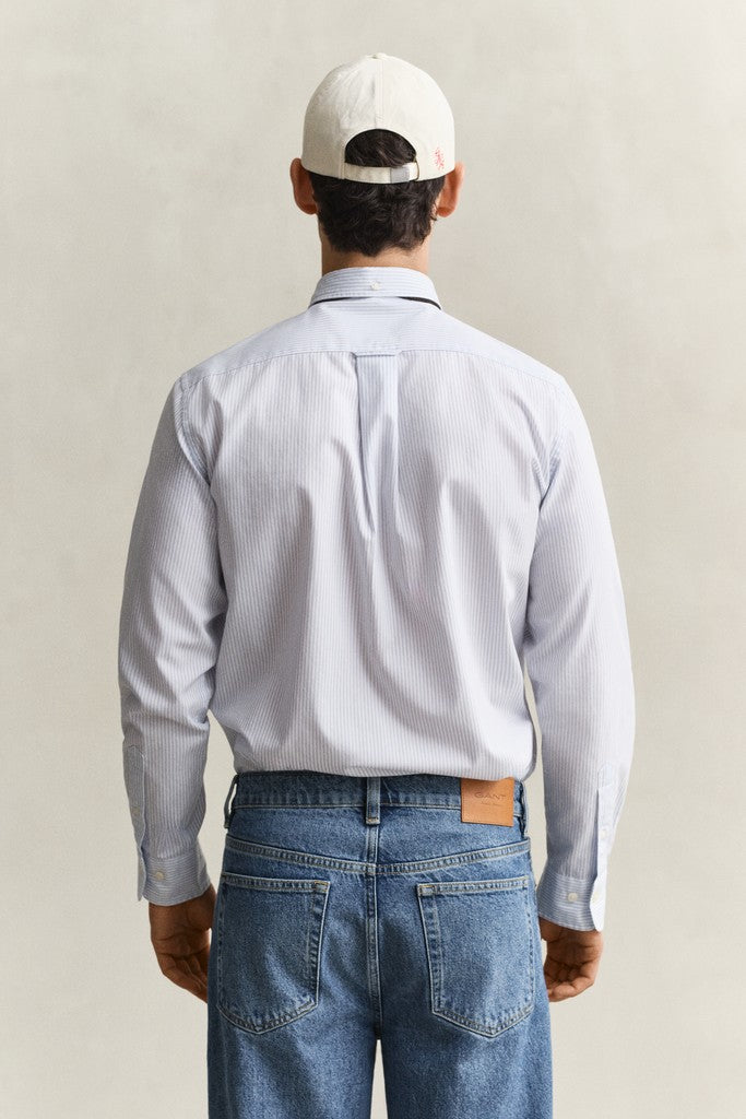 A man viewed from behind wearing a light blue long-sleeve button-down shirt, blue jeans, and a white baseball cap, set against a neutral beige background.
