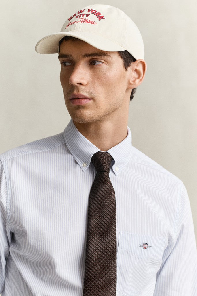 A man wearing a light blue and white striped GANT shirt, a dark brown tie, and a cream-colored baseball cap with red embroidered text. The shirt features a small embroidered shield logo on the chest pocket.