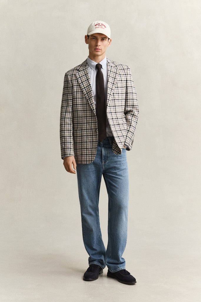 A man modeling a checkered blazer with blue jeans, a light stripe shirt, a dark tie, and a white baseball cap against a neutral studio background.