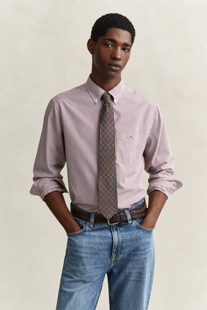 A man modeling a light red and white striped long-sleeve dress shirt with a patterned brown, gold, and blue tie, paired with blue jeans and a brown belt, against a neutral beige background.