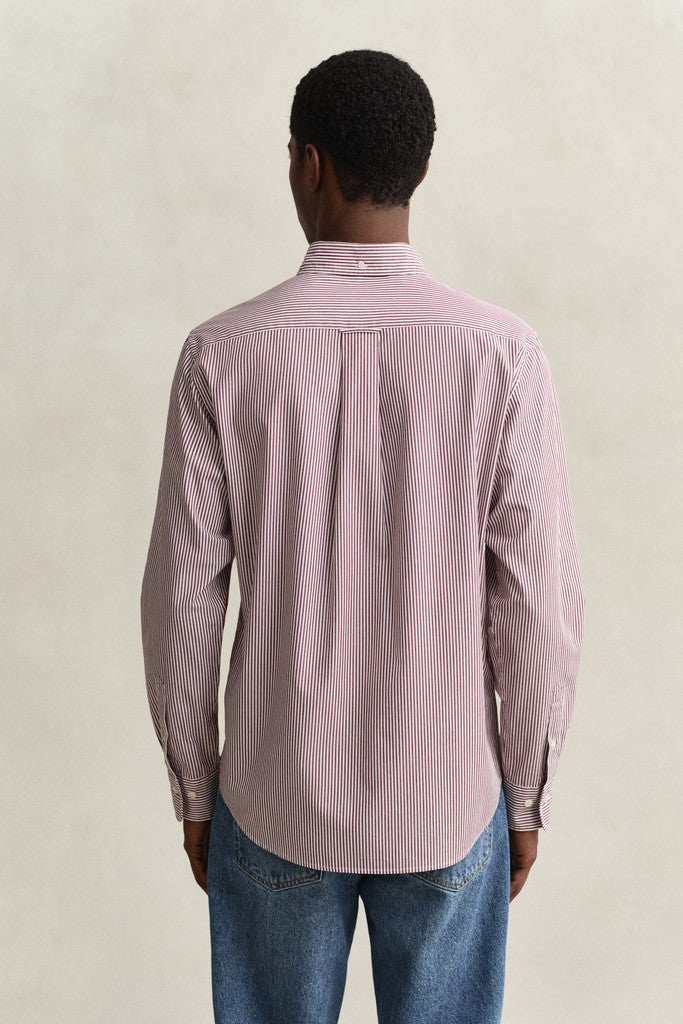 A man viewed from behind wearing a light red and white vertically striped button-down shirt and blue jeans, standing against a neutral beige background.